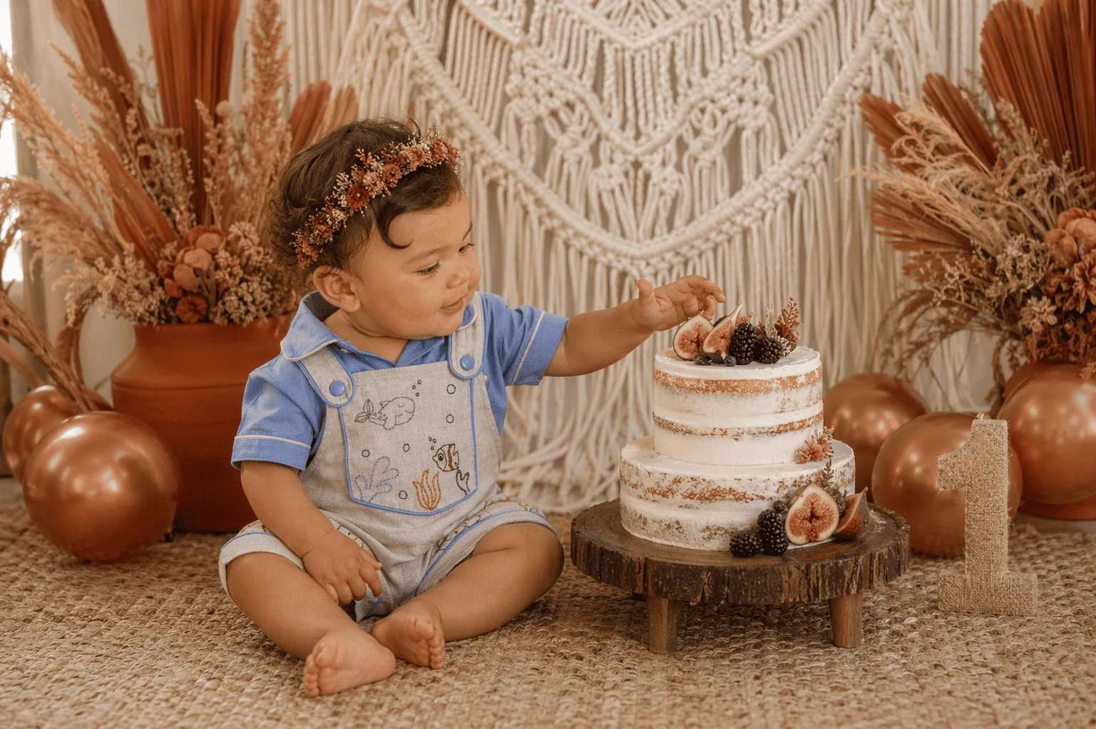 Bebê menina de 1 ano com coroinha de flores secas sentada no chão estendendo a mão pra bolo de 2 andares com figos e amoras sobre base de madeira rústica — decoração de mesversário/aniversário com macramê, balões metalizados cobre, flores secas em tons ferrugem · vestindo Jardineira Premium Fundo do Mar · cenário de festa sofisticada