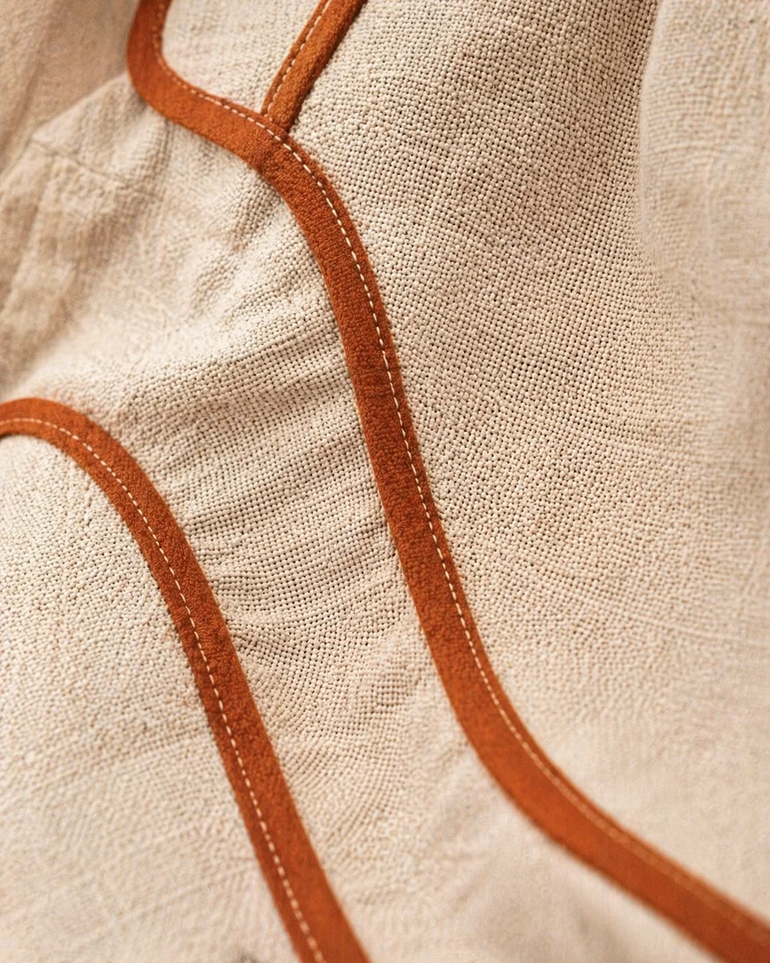 Macro do avesso interno da Jardineira Premium em linho areia — três tiras de viés caramelo-terracota costuradas curvando elegantemente cobrindo todas as costuras internas, pesponto duplo cream contrastante visível, zero fio solto · macro suave editorial Portra 400