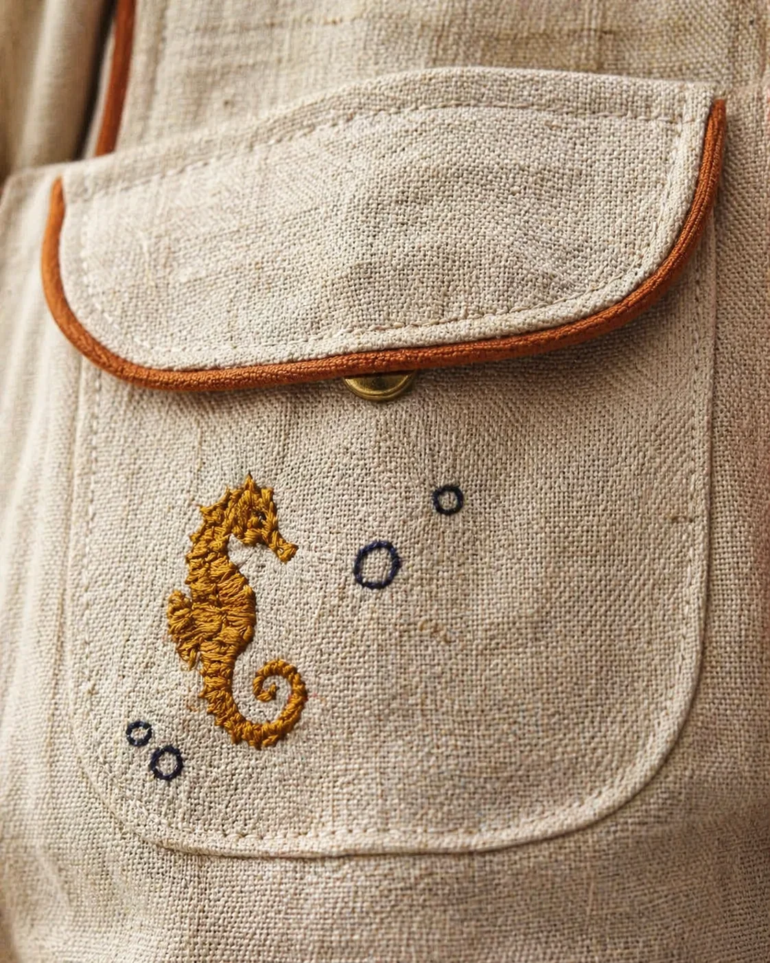 Macro do bolso frontal da Jardineira Premium em linho natural cor areia — bordado de cavalo-marinho em fio dourado-mostarda, bolhas pontilhadas em azul-marinho, viés caramelo-terracota contornando a aba, botão de pressão bronze pequeno aparecendo na borda · macro Kinfolk editorial Portra 400