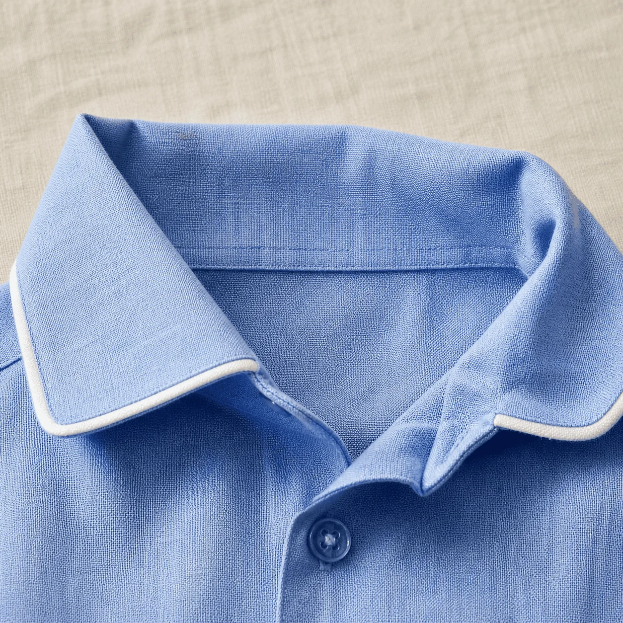 Macro da gola padre da Camisa Social em cetim azul céu · vivo branco contornando o colarinho · botão central · acabamento de camisa social adulto numa peça infantil.