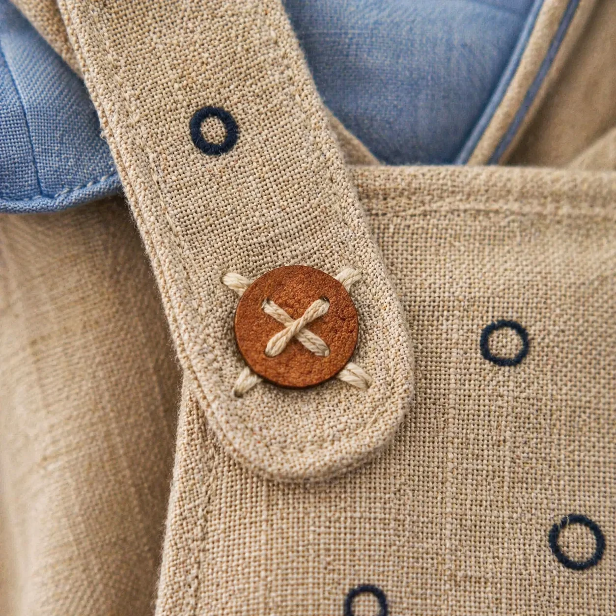 Macro do botão de couro caramelo costurado em ponto cruzado X em linho natural areia — junção da alça da Jardineira Premium com bordado pontilhado azul-marinho ao redor, sliver de camisa azul chambray no topo · macro tactile Kinfolk Portra 400